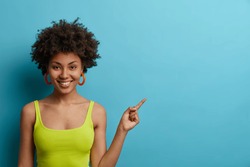 Friendly looking positive young woman gives suggestion or recommendation, points index finger on copy space, dressed in bright summer outfit, isolated on blue background. People and advertisement