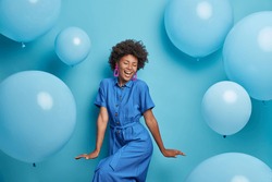 Carefree joyful curly woman dances happily, dressed in blue dress, chills at party around inflated helium balloons, feels playful, enjoys favorite holiday, has upbeat festive mood. Moment of joy