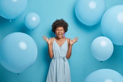 Shot of upbeat cheerful dark skinned woman feels very happy and excited, raises palms and laughs, spends free time on party, wears nice blue summer dress with earrings and rings, poses near balloons