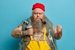 Strict bearded annoyed fisherman clenches fist angrily, looks with frowned face, uses fishing nets, keeps smoking pipe in mouth, wears yellow overalls, poses against blue background, works on boat