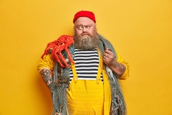 Serious thoughtful fisherman with thick beard, caught big octopus and smokes pipe indoor. Old experienced sailor poses on fishing boat against yellow background. Pensive captain has marine adventure