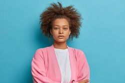 Portrait of serious dark skinned woman with combed curly bushy hair has natural beauty looks directly at camera wears pink jumper isolated on blue background. Human face expressions concept.