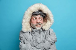 Photo of European man trembles from cold after going skateboarding crosses hands over body tries to warm himself wears grey winter jacker with fur hood and gloves has frozen face covered by ice
