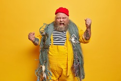 Annoyed emotional male sailor or professional seaman has sea cruise poses with fishing net raises tattooed arms and yells outraged wears red hat and yellow overalls stands indoor. Marine life concept