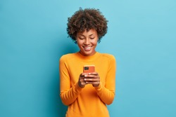 Online lifestyle concept. Cheerful good looking woman with Afro hair sends text messages via mobile phone dressed casually searches gifts for holiday in internet uses smartphone app browses webpage