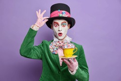 Photo of shocked mad hatter being on tea party wears big hat lace gloves and green costume poses with beverage has professional bright makeup isolated on purple background. Halloween carnival