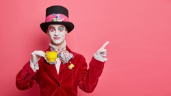 Smiling glad hatter in fashionable costume has aristocratic manners drinks tea and indicates at copy space isolated over rosy background. Man has image of classic tale character for halloween