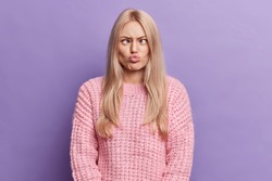 Funny blonde young woman has silly grimace crosses eyes and pouts lips makes brainless face wears knitted sweater isolated over purple background foolishes around. Human face expressions concept