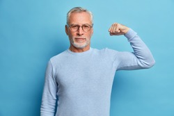 Senior bearded man shows muscles after practising bodybuilding wears transparent glasses and basic jumper poses against blue studio background. Look I am very strong and healthy. Perfect biceps