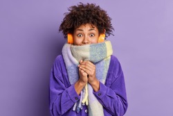 Afro American woman feels very cold during freezing weather wears purple jacket and warm scarf around neck wals on street during winter listens music via wireless headphones keeps hands together