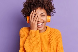 Close up portrait of overjoyed dark skinned woman keeps hand on face and smiles carefree listens favorite music in headphones dressed casually expresses positive emotions isolated on purple wall