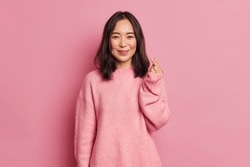 Pretty dark haired woman with gentle smile makes korean like sign expresses love to you wears casual long sleeved jumper isolated over pink background. Mini heart gesture. Body language concept