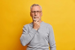Senior thougthful man holds chin and looks pensively aside makes plannings wears spectacles and casual grey jumper isolated over vivid yellow background. Handsome grandfather considers something