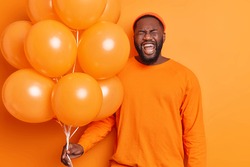 Overjoyed bearded man laughs positively has fun during birthday party holds bunch of balloons wears hat and sweater isolated over vivid orange background. People holiday celebration concept.