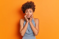 Nervous worried Afro American woman clenches teeth looks embarrassed at camera wears fashiobnable outfit afraids of bad consquences poses in studio against orange background. Scared female model