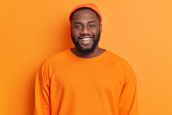 Portrait of cheerful bearded African American man has happy expression smiles broadly has white perfect teeth wears hat and sweater isolated over orange background. Positive emotions concept