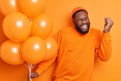 Overjoyed male student celebrates success makes yes gesture being on graduation party holds inflated balloons dressed in casual jumper isolated over orange background. People happiness concept