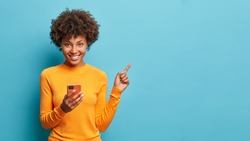 Horizontal shot of happy dark skinned Afro American woman enjoys mobile communication and modern technologies poses against blue background points away on free space for your advertising content