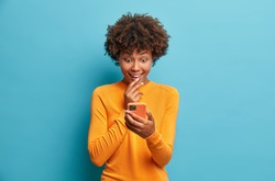 Happy surprised Afro American woman browses internet and networking in social media tests new application for smartphone wears casual jumper isolated over blue background. Mobile communication