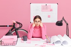 Professional tired female architect works on building sketch feels tired of long hours working checks graphic outlay tries to improve idea of planning poses at desktop surrounded with papers stickers
