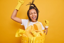 Happy woman housemaid dances carefree keeps arms raised poses near laundry basin has fun while doing household chores sings song along isolated over vivid yellow background. Housekeeping concept
