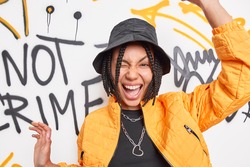 Urban style lifestyle youth and emotions concept. Positive carefree hipster girl has dreadlocks raises arms winks eyes wears black hat and fashionable orange jacket poses against graffiti wall.