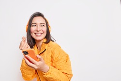 Studio shot of happy Asian millennial girl makes eastern like sign smiles gladfully looks away dressed in orange jacket uses smartphone and headphones for listening favorite music poses indoor