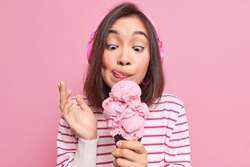 Asian brunette young woman looks at appetizing ice cream licks lips eats yummy frozen dessert listens music via headphones dressed in striped jumper isolated over pink background. Mmm how tasty