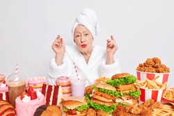 Lovely wrinkled lady wears red lipstick enjoys cheat meal day surrounded by junk food eats tasty snacks isolated over white background cosumes large portions isolated over white studio background