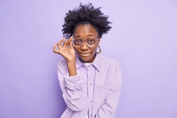 Portrait of dark skinned woman looks attentively through spectacles wears stylish jacket listens information attentively poses against purple background. Monochrome shot. Human face expressions