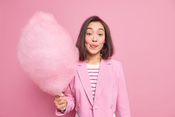 Good looking brunette Asian woman licks lips as holds appetizing sweet candy floss dressed in fashionable formal outfit isolated over pink background. Teenage girl with delicious cotton candy