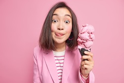 Lovely Asian woman with dark hair licks lips starves to eat ice cream likes sweet junk food dressed formally isolated on pink background. Summer time concept. Pretty female model likes frozen dessert