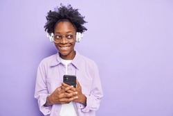 Positive dark skinned curly woman holds mobile phone downloads songs to her playlist wears wireless headphones big spectacles fashionable jacket isolated over purple background with copy space