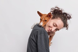 Sideways shot of adult girl carries puppy which embraces her around neck walkk together isolated over white background. Female dog lover poses with pet looks away likes animals. Care and love