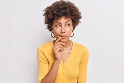 Horizontal shot of thoughtful young African American woman looks aside has dreamy expression interesting idea in mind wears casual yellow jumper isolated over white background makes decision