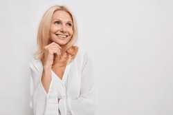 Beautiful young European woman of middle age smiles gently keeps hand under chin looks away with dreamy expression wears silk blouse isolated over white background copy space for advertisement