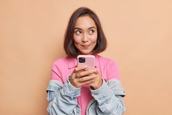 Dreamy young pretty Asian woman with dark hair holds mobile phone has thoughtful expression thinks about received message wears pink t shirt denim jacket looks away isolated over beige background.