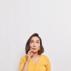 Vertical shot of dreamy thoughtful young Asian woman with dark hair focused above considers something wears casual yellow jumper isolated over white background copy space for your advertisement