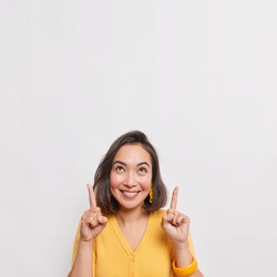 Positive brunette Asian woman points above shows place for your advertising text smiles pleasantly wears yellow jumper earrings and bracelet isolated over white background. Great idea cool offer