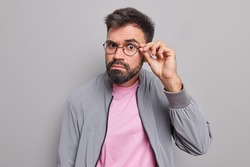 Serious bearded man keeps hand on rim of spectacles has attentive gaze directly at camera has surprised expression wears jacket isolated over grey background listens something. Monochrome shot