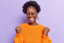 Joyful African American woman makes yes gesture feels like winner smiles broadly celebrates win feels excited keeps eyes closed wears casual orange knitted sweater isolated over purple background