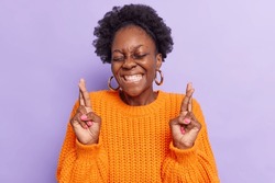 Positive dark skinned curly young woman smiles broadly makes wish prays with good attitude waits for dream come true keeps eyes closed wears knitted orange jumper isolated over purple background