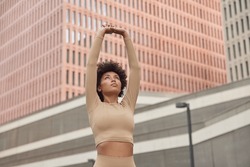 Motivated American woman raises arms over herself dressed in sportswear stretches before workout poses against cityscrapers outside has pilates exercises. Healthy lifestyle and activity concept