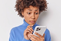 Close up shot of puzzled curly haired woman looks at smartphone screen with concerned expression reads bad news wears casual blue turtleneck isolated over white background has complicated life
