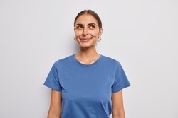 Portrait of dreamy dark haired woman smiles gently concentrated above thinks about something very pleasant recalls nice memories wears casual blue t shirt isolated over white studio background