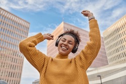 Positive carefree curly haired woman dances with joy enjoys favorite audio track via headphones dressed in casual knitted jumper poses against skyscrapers and blue sky spends free time at city