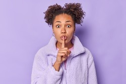 Cute shocked young woman makes silence gesture presses index finger over lips shows hush sign says shh dressed in outerwear spreads gossips isolated over purple background. Body language concept