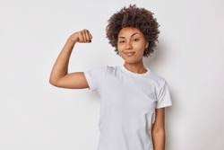 Confident woman feels strength and power raises arm flexes biceps and looks proud of her own achievements has strong muscles wears casual t shirt isolated over white background brags with fit body