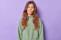 Studio shot of displeased bored woman purses lips and looks with uninterested expression wears casual jumper poses alone against purple background. Upset gloomy teenage girl tired of distance studying
