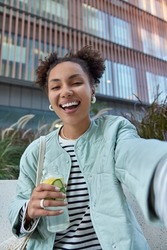 Happy optimistic girl with two hair buns dressed in jacket enjoys free time and walking in city holds bottle of detox drink makes selfie poses against modern building has fun during daytime.